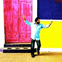 A smiling man in a turban poses cheerfully against a vibrant pink door and yellow and purple walls.