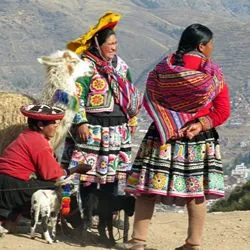 Indigenous Andean women wearing vibrant, traditionally embroidered clothing and floral skirts, standing with a fluffy, decorated llama against a mountainous background.