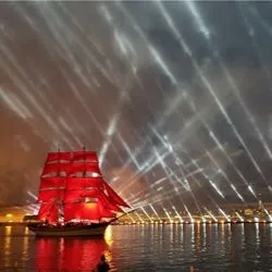 A tall ship with glowing scarlet sails glides down a river at night, illuminated by a dramatic display of white spotlights beaming into the sky.