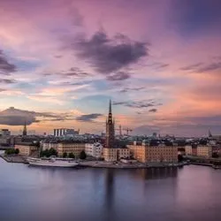 Panoramic twilight view of the Stockholm, Sweden waterfront and the Riddarholmen island skyline under a pink and orange sunset sky.
