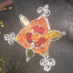A traditional kolam or rangoli design outlined in white powder and filled with colorful flower petals on a dark floor.