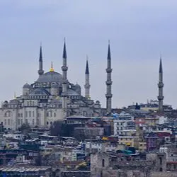 Panoramic view of the historic Sultan Ahmed Mosque (Blue Mosque) in Istanbul, Turkey, rising above the surrounding city buildings under a cloudy sky.
