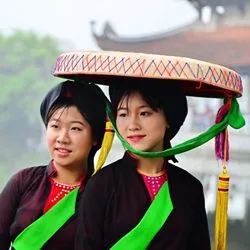 Two women wearing traditional Vietnamese garments and headwear standing outdoors against a blurred, foggy background.