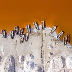 Top-down aerial photograph of traditional wooden boats lined up on the sandy shoreline of Lake Retba (Lac Rose) in Senegal, contrasting against the bright orange-pink water.