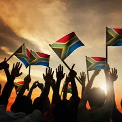 Silhouetted hands waving South African flags against a sunset sky.