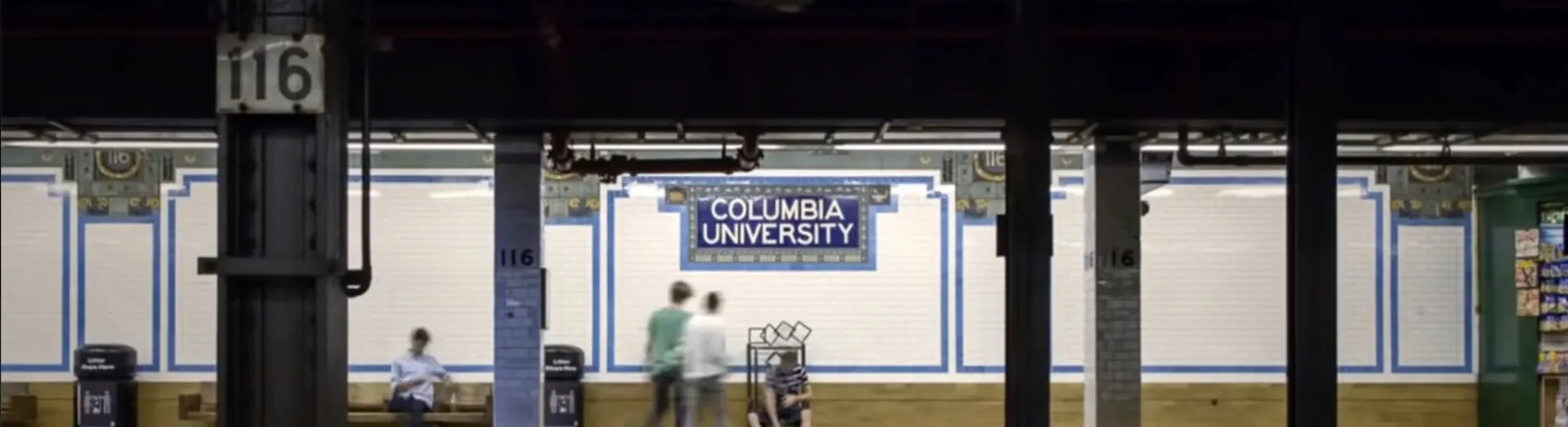 NYC subway platform at the 116th Street–Columbia University station, featuring a blue "COLUMBIA UNIVERSITY" mosaic sign and white tiled walls.