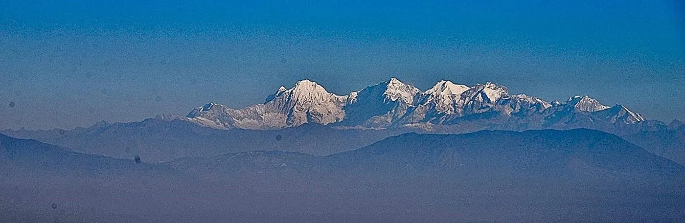 A panoramic view of a snow-capped mountain range rising above hazy, dark blue foothills under a clear blue sky.