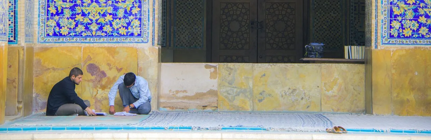 Two young men sit on rugs in an outdoor courtyard of a building adorned with intricate blue and yellow floral mosaic tiles, focusing on open books in front of them.