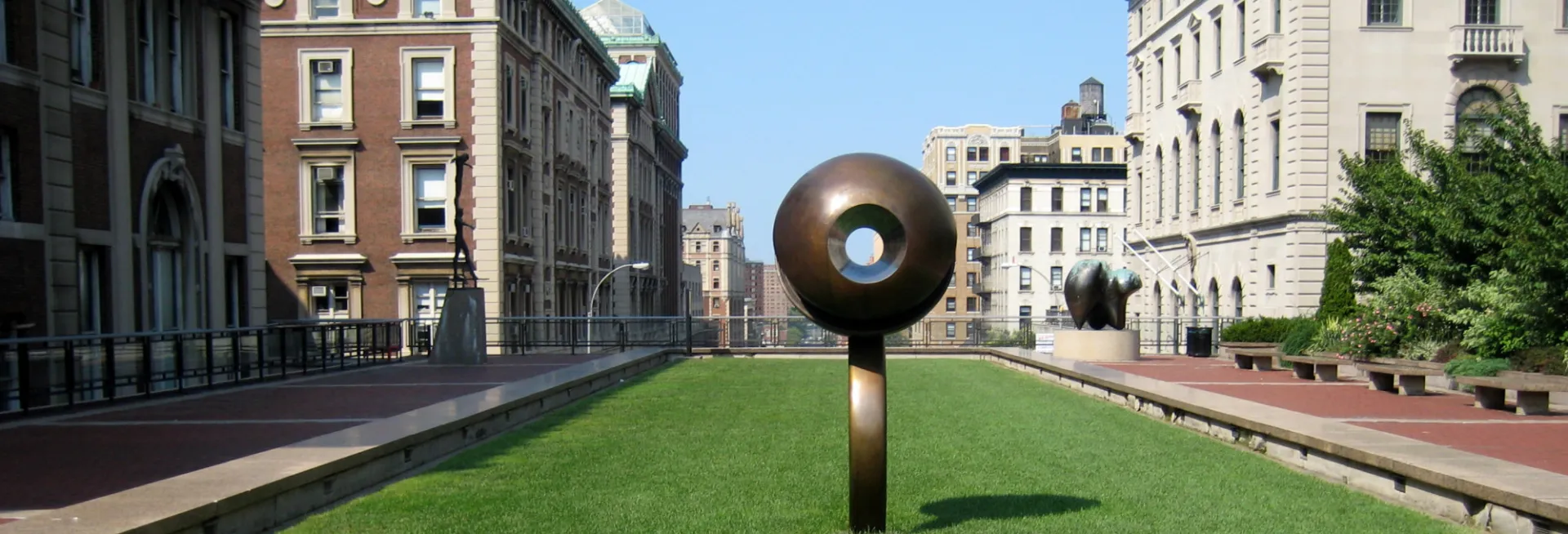 Elevated grassy lawn at Revson Plaza on Columbia University campus, featuring a large bronze sculpture and surrounding university buildings.