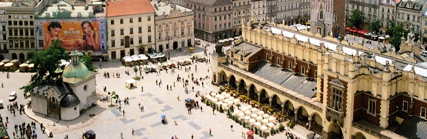 An aerial view of Krakow's Main Market Square in Poland, showing the historic Cloth Hall, St. Adalbert's Church, and pedestrians walking across the sunny plaza.