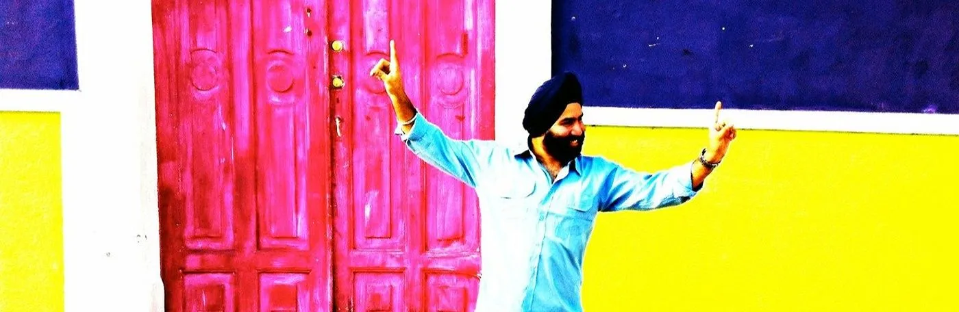 A smiling man in a turban poses cheerfully against a vibrant pink door and yellow and purple walls.