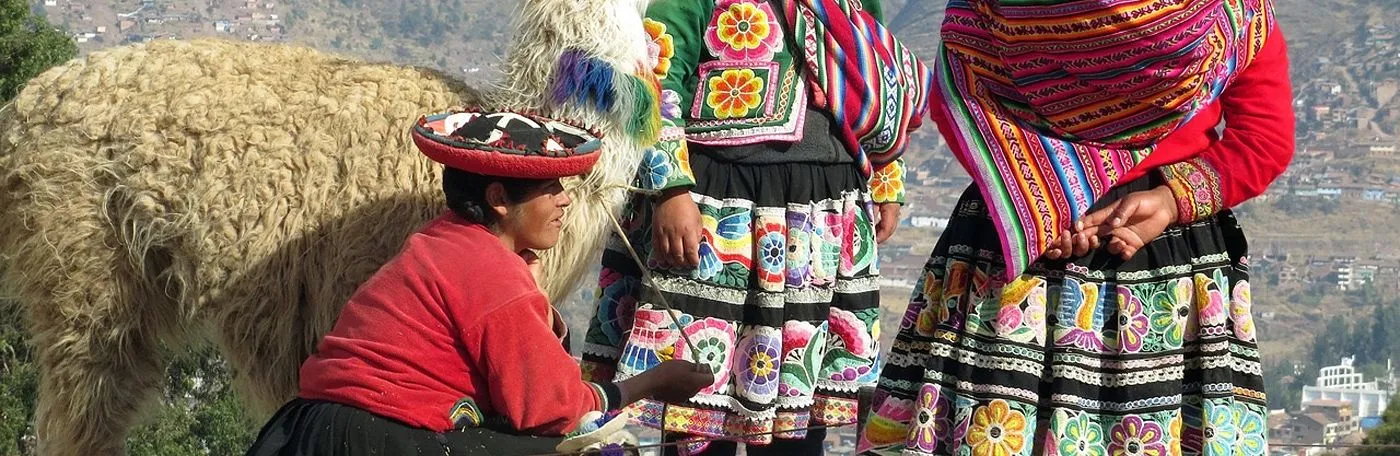 Indigenous Andean women wearing vibrant, traditionally embroidered clothing and floral skirts, standing with a fluffy, decorated llama against a mountainous background.
