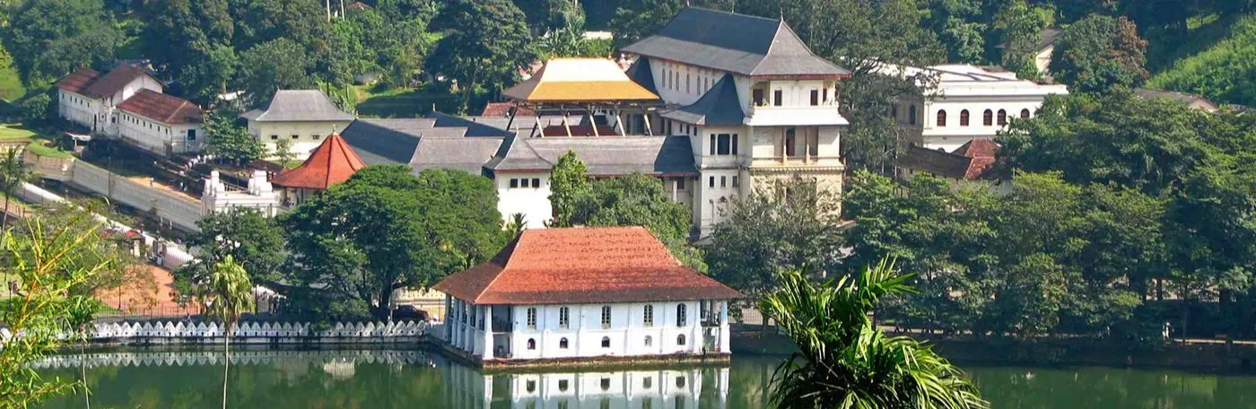 An elevated view of the Temple of the Tooth complex in Kandy, Sri Lanka, featuring traditional white buildings with tiled roofs situated next to a calm lake, surrounded by lush green trees.